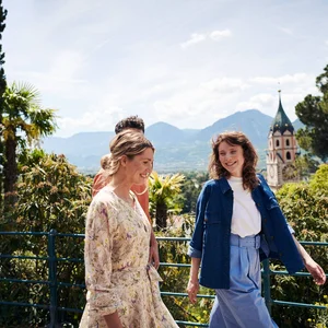 Drei Menschen gehen draußen mit Berg- und Kirchenturmkulisse im Hintergrund spazieren