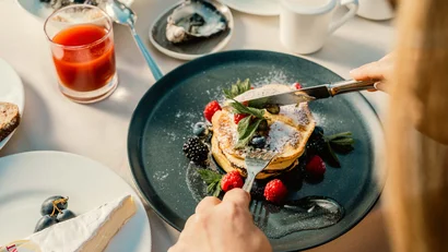 Person schneidet Pancakes mit Beeren und Puderzucker auf blauem Teller