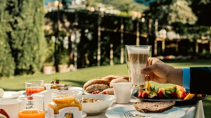 Hand hält Latte Macchiato bei Frühstück mit Brot und Obst im Garten