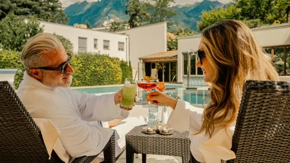 Zwei Personen im Bademantel stoßen am Pool mit Cocktails an