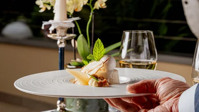 Servierer reicht kunstvoll angerichtetes Dessert auf weißem Tisch mit Kerze und Glas