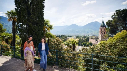 Drei Menschen spazieren in sonniger Berglandschaft mit historischen Gebäuden