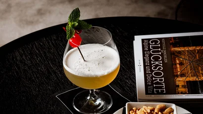 Cocktail mit Minze und Kirsche neben einer Schüssel Erdnüsse und einer Zeitung